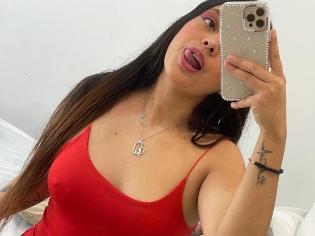 Violetta Rouse, com longos cabelos castanhos, língua de fora, vestindo uma blusa vermelha de alças finas, mostrando os seios, segurando um celular com uma capa brilhante, usando um colar com um coração e com uma tatuagem no pulso direito..