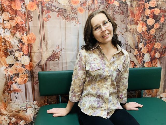 DaisyEdit with glasses, smiling, wearing a floral button-up shirt and black pants, sitting on a green chair, hands on her ass. Her tits are small and she's cute.