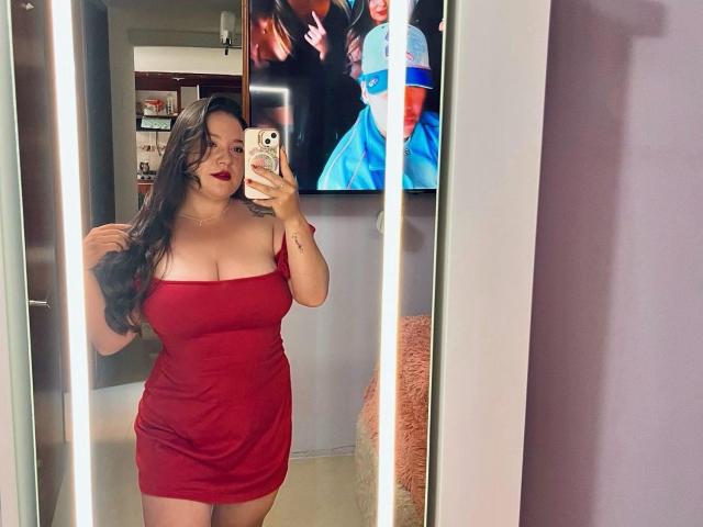 EmiliaMora with big tits in a tight red dress, taking a mirror selfie, holding her long dark hair, red lipstick, standing in a room with a TV behind her.
