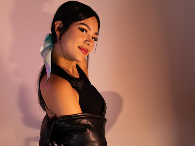 ValerieCasanova with dark hair, wearing a black tank top and leather jacket, showing off her tits with a cute smile, a shiny blue ribbon in her hair.