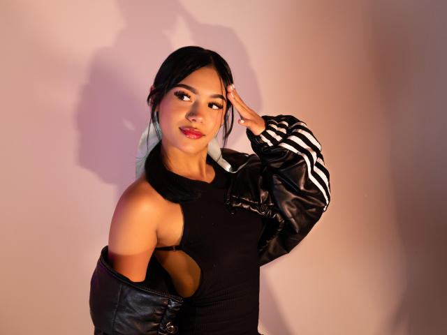 ValerieCasanova with black hair, wearing a black cut-out dress and black leather jacket with striped sleeves, posing with hand on head, smiling, red lipstick, white earrings, sexy.