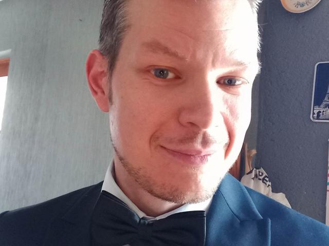 TheLoveMen69, a bald Caucasian man with light skin and blue eyes, wearing a black suit with a bow tie, smiling slightly with a subtle beard and stubble.