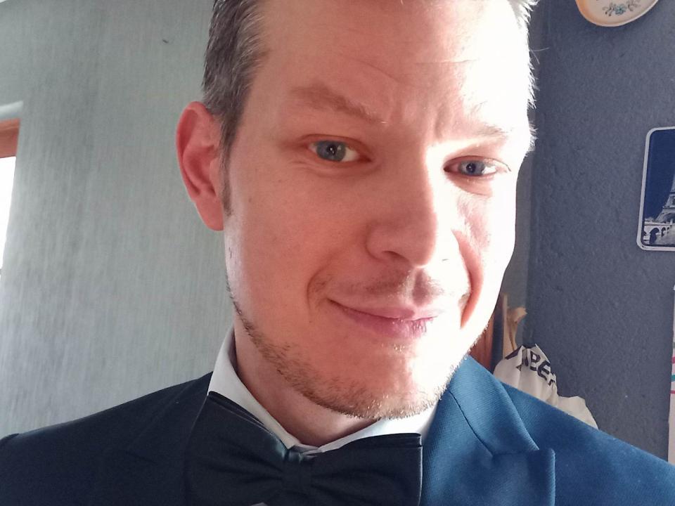 TheLoveMen69, a bald Caucasian man with light skin and blue eyes, wearing a black suit with a bow tie, smiling slightly with a subtle beard and stubble.
