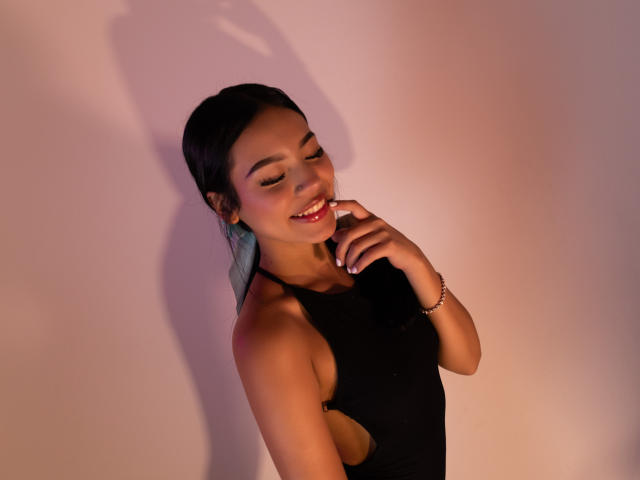  ValerieCasanova with dark hair, smiling, wearing a black halter top, touching her chin, showing her small ass, bracelet on her wrist, shadow on the wall.