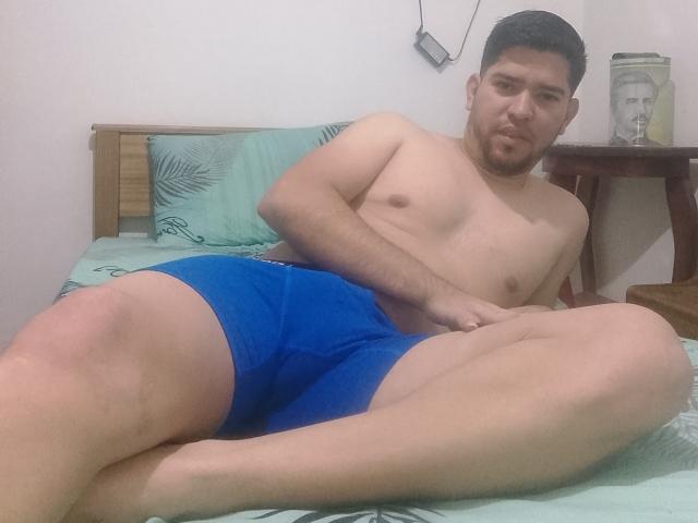 ChristopherRusso is shirtless, wearing blue boxer shorts, lying on a bed with a green pillow, legs crossed, hand on his crotch, short dark hair, and a neutral expression.