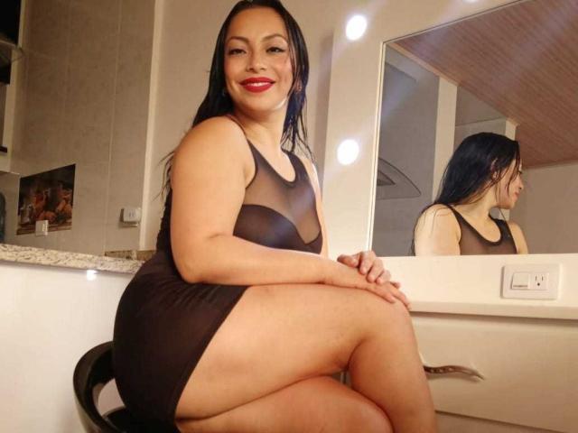 AnayPorter, a curvy Latina woman with long black hair, red lipstick, and dark sheer dress, sitting on a chair, showing her ass and reflection in a mirror.