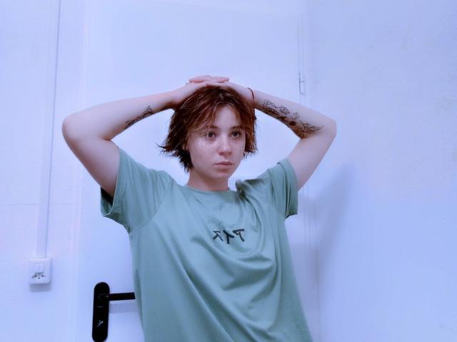 AnnaWestt with arms raised, wearing a green T-shirt, has short red hair and tattoos on both arms. She's standing in a white room, looking serious.
