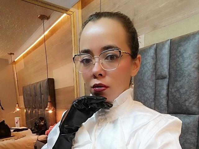 BarbaraLeythonn with glasses, red lipstick, black gloves, and white shirt, holding her chin, looking at camera, in a modern bedroom.
