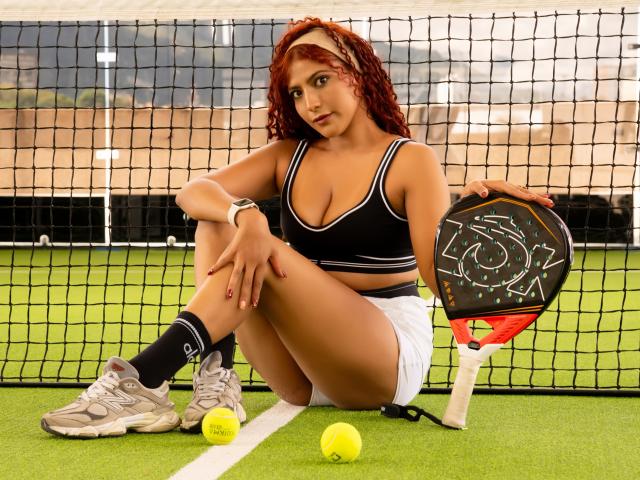 Mia Bianchi, con capelli rossi ricci, reggiseno sportivo nero, pantaloncini bianchi e calze nere, seduta su un campo da tennis, che tiene una racchetta, palle da tennis e mostra il sedere..
