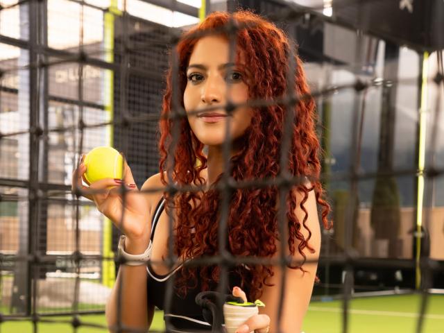 MiaBianchi with curly red hair, holding a yellow tennis ball, smiling, wearing a black sports top, in front of a tennis net. Her tits are small, she's cute.