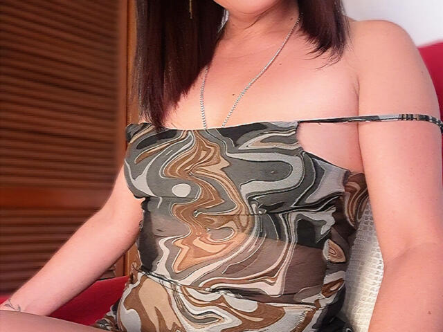 DhannaRose with medium tits, wearing a strapless, marbled dress, silver necklace, brown hair, wooden background, red chair, and a subtle tattoo on her right wrist.