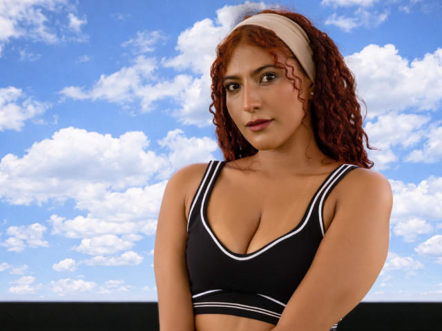 Mia Bianchi, con i capelli rossi ricci, la pelle abbronzata e una fascia bianca tra i capelli, indossa un reggiseno sportivo nero con bordi bianchi, mostrando il suo seno, di dimensioni piccole o medie, contro un cielo blu..