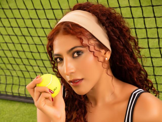 MiaBianchi with curly red hair, beige headband, holding yellow tennis ball, black-and-white striped top, green grass, black net, looking at camera, medium skin, red nail polish, earrings.