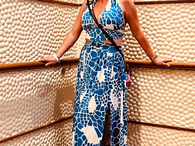 Zuckerschnecke-hot wears a blue and white abstract patterned dress with a high slit, revealing a toned midsection. She leans on a wooden railing with her hands, showcasing a silver bracelet on her right wrist. The dress has a halter neck and a black strap crosses her chest. The textured background is a wall of white, pebble-like shapes. The image is brightly lit, emphasizing the vivid blue and white colors of the dress.