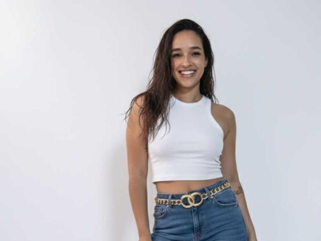 AlissonCooper with wet brown hair, white sleeveless crop top, blue jeans with gold chain belt, smiling, standing against white wall, hands by sides, small tattoo on right arm.