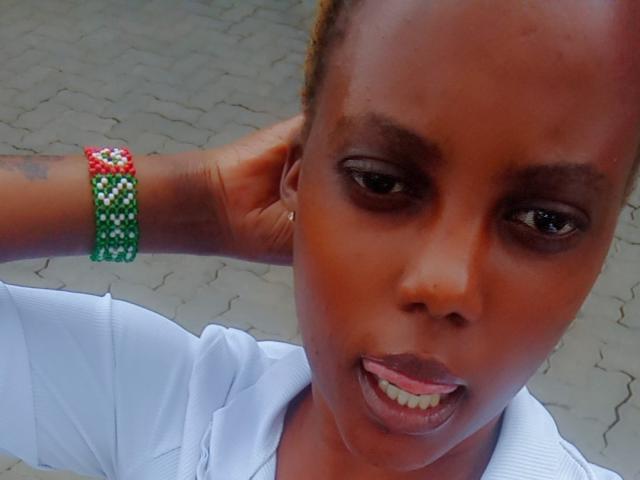 Starlesty has a dark brown face, short hair, green and red beaded bracelet, white shirt, and slightly parted lips with a tongue showing. Her hand is behind her head.