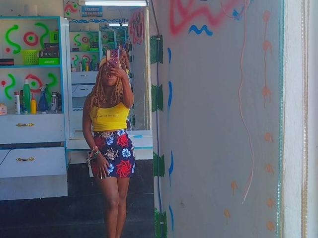 Starlesty with long blonde hair, yellow tank top, and colorful floral skirt, standing in front of a mirror, holding phone, in a brightly decorated room with graffiti.