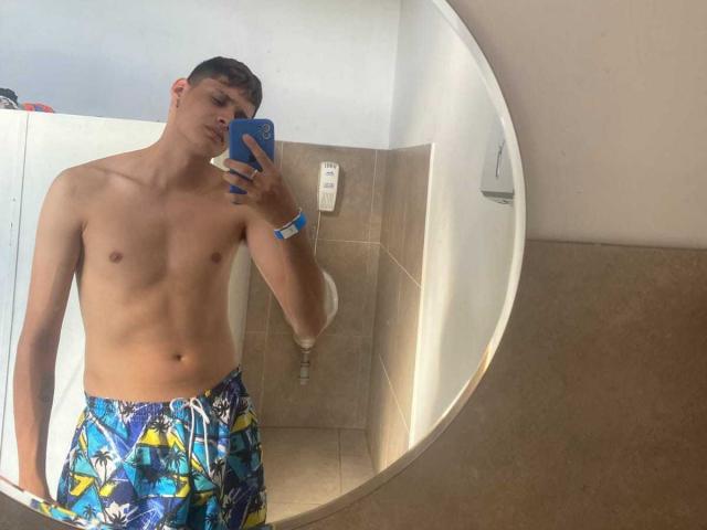 ElFlaquitoo, shirtless with blue and yellow swim trunks, holding a blue phone, mirror selfie showing a lean, smooth-chested, slim build, short brown hair, tan skin, in a bathroom.