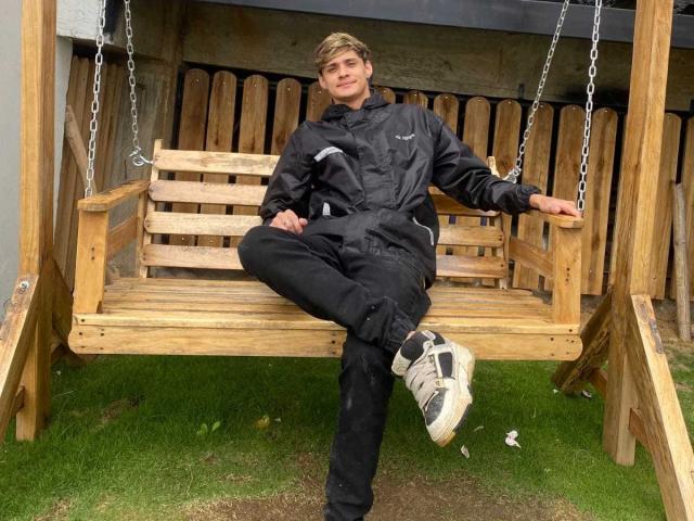 ElFlaquitoo is sitting on a wooden swing, wearing black clothes and white sneakers, with one leg raised and a relaxed smile. His blond hair is slightly messy.
