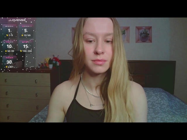 AliceGly with long blonde hair, pink lips, black halter top, necklace, and slight smile, in a bedroom. Her tits are small and she's looking down.