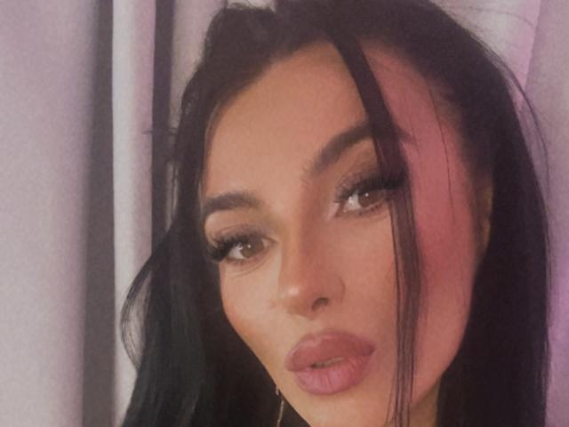 KinkyAmelie has dark, wavy hair, pinkish highlights, and full pink lips. Her brown eyes are framed by thick, long lashes, and she's wearing subtle makeup.