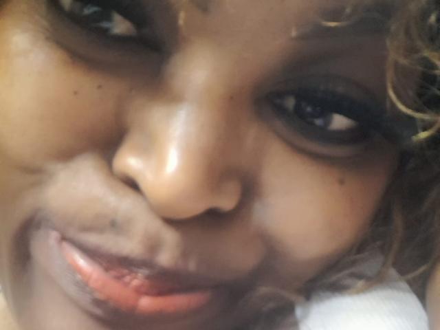 BigAssQueen's face is in the shot, smiling with her lips slightly parted, showing her teeth, and her curly brown hair is visible. Her dark eyes are looking to the side.
