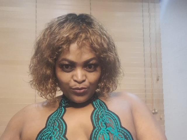 BigAsssQueen with curly brown hair, wearing a teal lace halter top, staring into the camera with a serious expression, showing her medium-sized tits.