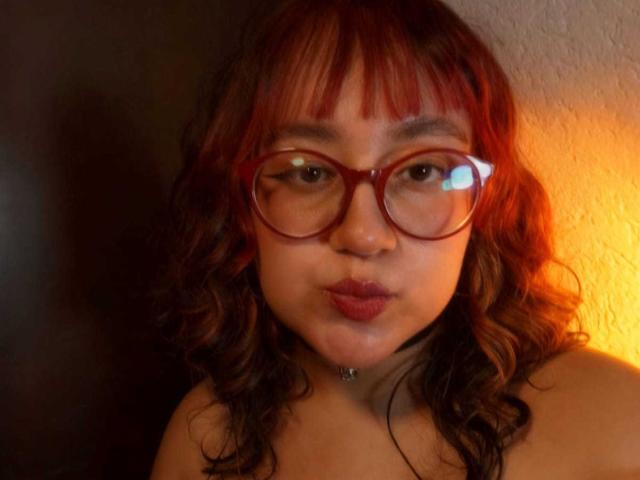NamiNerona with red curly hair, red glasses, and pouty red lips, showing her chubby ass with a black strap, wearing a black choker. Warm light in the background.