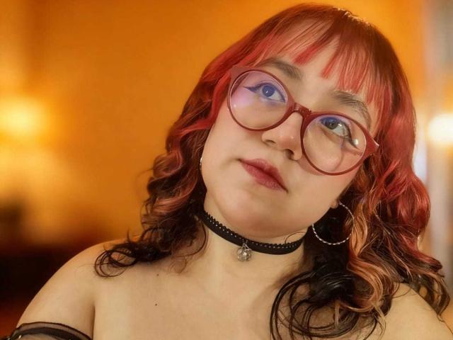 NamiNerona has red and black curly hair, wears red glasses, a black choker, and hoop earrings, with blue eyes and a neutral expression, showing her bare shoulders.