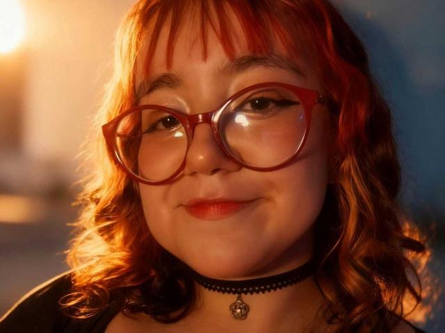 NamiNerona with red glasses, red and orange wavy hair, black choker, and subtle smile, illuminated by warm light, showing soft skin and red lips.