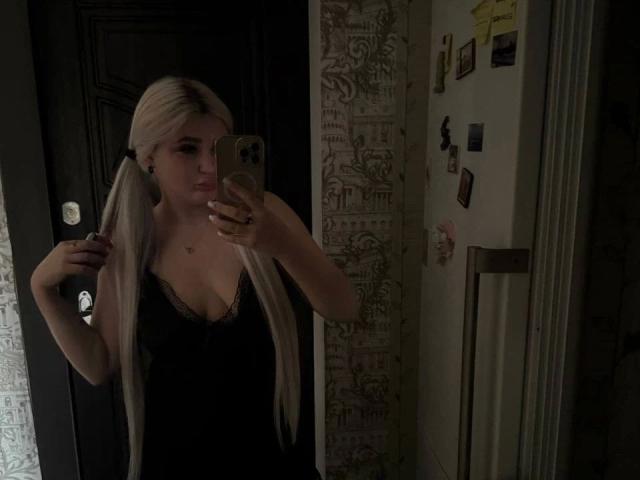 SlankyMic-hot with long platinum blonde pigtails, wearing a black lace-trimmed top, holding a phone, taking a mirror selfie, showing her small tits, in a dimly lit room.