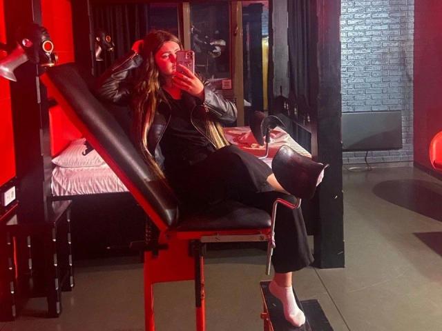 SlankyMic-hot is seated on a black leather and metal BDSM chair, taking a selfie with her right hand while resting her left on her head. She has long, wavy brown hair and wears a black leather jacket, black pants, and white socks. The room has red walls, a bed with white sheets, and a brick wall in the background. The lighting is red and moody.