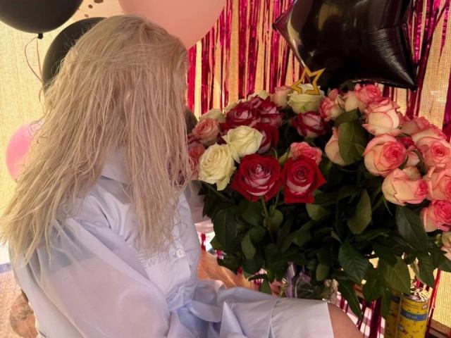 A photograph of SlankyMic-hot with wavy, blonde hair, wearing a light blue, button-up shirt. SlankyMic-hot is seated, facing a large bouquet of roses. The bouquet has a mix of red, white, and pink roses with green leaves. The background features pink and gold striped curtains. Black balloons are partially visible. The bouquet is to the right, and SlankyMic-hot is slightly turned towards it. The image has a warm, indoor lighting.