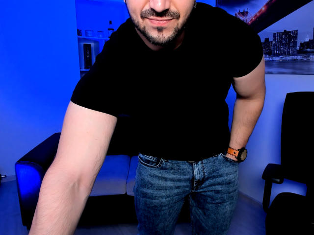 JaceThorne with a muscular build, black t-shirt, and blue jeans, leaning forward, showing his arm, with a beard and mustache, wearing a wristwatch, in a blue-lit room.