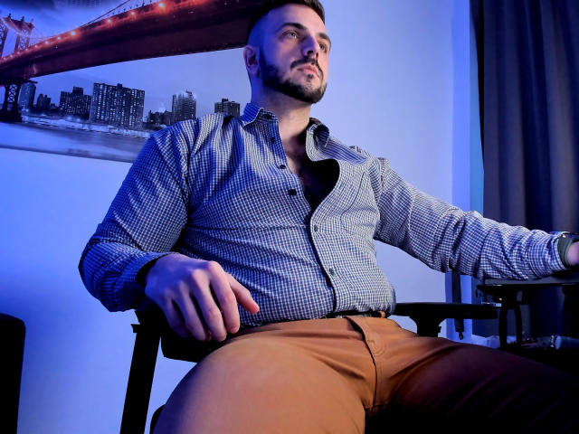 JaceThorne, muscular with a beard, wearing a blue checkered shirt and brown pants, sits in a chair, looking up, with a red bridge and city skyline in the background.