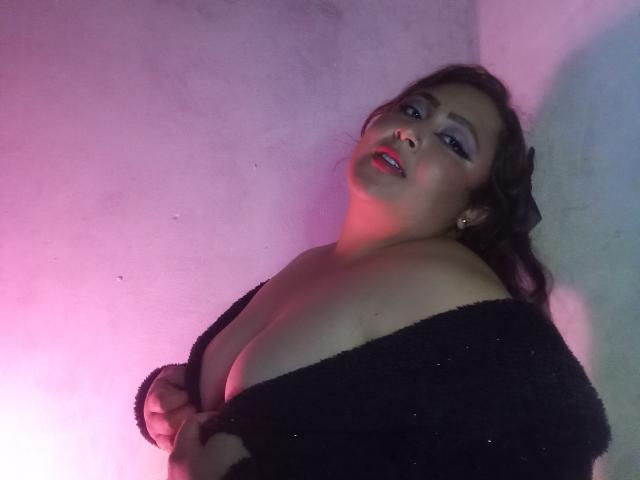 ValeriaGotOne with her tits slightly exposed, wearing a black off-shoulder sweater, has pink and purple lighting on her, looking up with red lipstick and dark wavy hair.