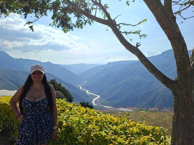 KatherinLatin stands in a sunlit, mountainous landscape wearing a blue floral dress, pink cap, and smiling, with a tree and green bushes in the foreground.