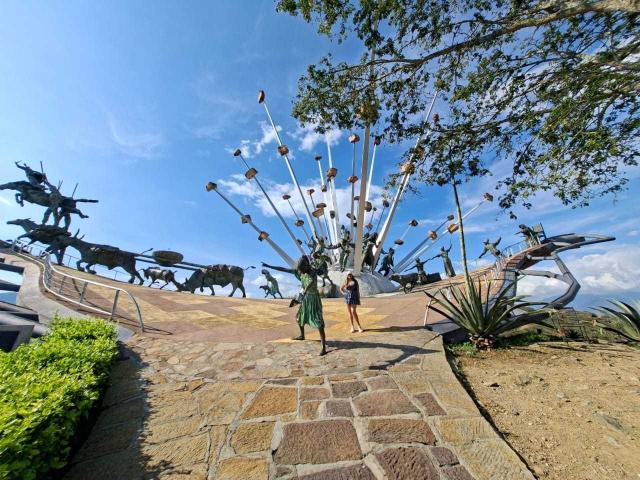 KatherinLatin si trova su un sentiero di pietra, fiancheggiata da enormi sculture meccaniche di uccelli e aeroplani, con una struttura alta, bianca e a forma di ventaglio sullo sfondo. Cielo azzurro limpido sopra..