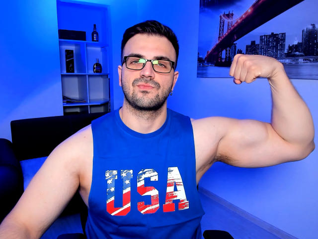 JaceThorne, muscular guy with short dark hair, glasses, beard, flexing right arm, wearing blue USA tank top, blue room, city wall art, bottle on shelf in background.