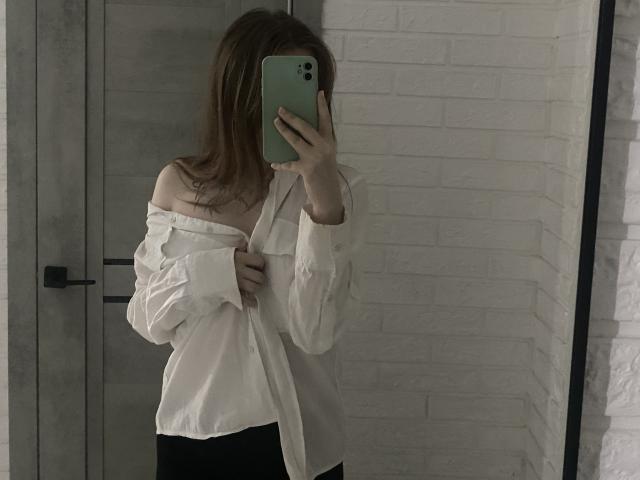 KirstieDevost taking a mirror selfie with her phone, wearing an off-shoulder white shirt, her tits partially exposed, standing in a white brick-walled room.