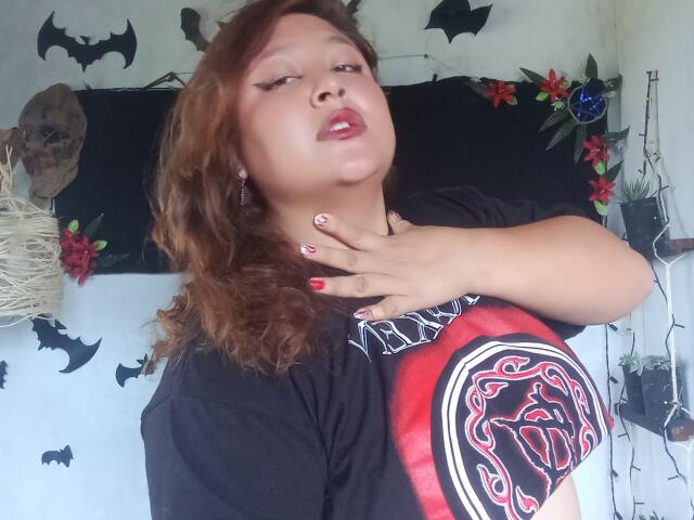 ValeriaGotOne with wavy brown hair, red lipstick, and black t-shirt with red and black graphic, hand on neck, nails painted red with white designs, looking slightly up.