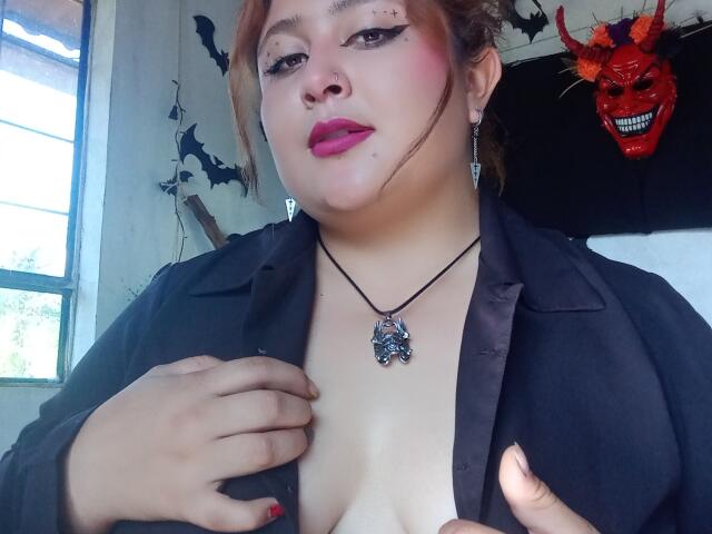 ValeriaGotOne with pink lipstick, black shirt open, showing her tits, holding shirt, necklace with dark gem, earrings, red nail polish, and a red devil mask in the background.