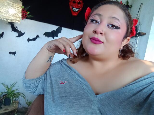 ValeriaGotOne with pink lipstick, gray off-shoulder top, red nail polish, and a tattoo on her right hand, posing with a red bow in her hair, looking at the camera.