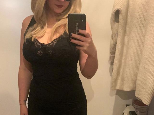 Elisa92-hot with blonde hair, wearing a black lace-trimmed dress, holding a phone in a mirror selfie, showing her tits and a small part of her ass.