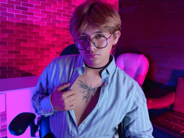 BenjiVampire with glasses, blonde hair, and a blue striped shirt, open to show a chest tattoo of a butterfly, wearing a black choker, sitting in a chair.