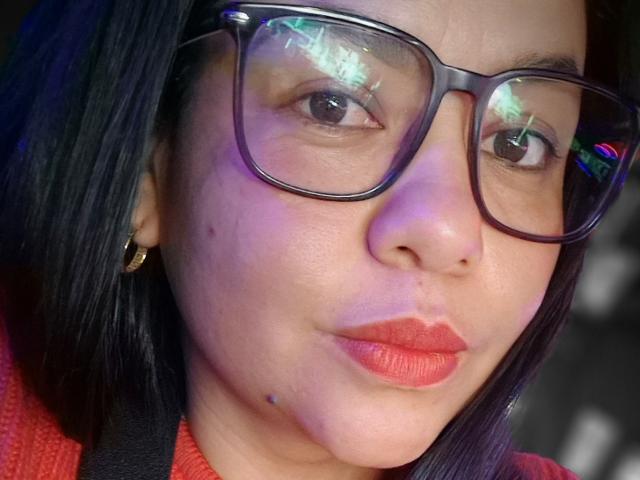 XBigTitxx has a puffy face with red lipstick, black glasses, and a small mole on her chin. She has dark hair and wears a red sweater.