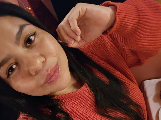 XBigTitxx has light brown skin, black hair, and wears a red rib-knit sweater, lying down with a slight smile, right hand near her face. Her brown eyes look at the camera.