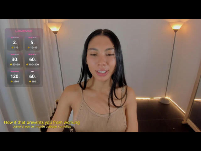 DayannaTecca with long black hair, tan tank top, small tits, and a cute smile, standing in a room with two lit floor lamps. She's looking down with a relaxed expression.