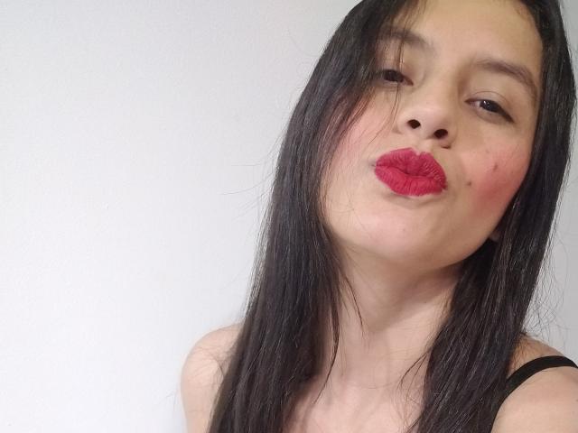 Madisson Fontaine, con cabello negro, lápiz labial rojo y un pequeño grano en la mejilla, frunciendo los labios. Lleva un top negro con tirantes y mira ligeramente hacia un lado..