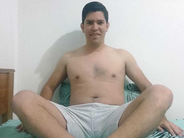 Shirtless ChristopherRusso, with short black hair, sitting on a bed, legs spread, wearing grey shorts, smiling, arms resting on green bedspread, white wall in background.